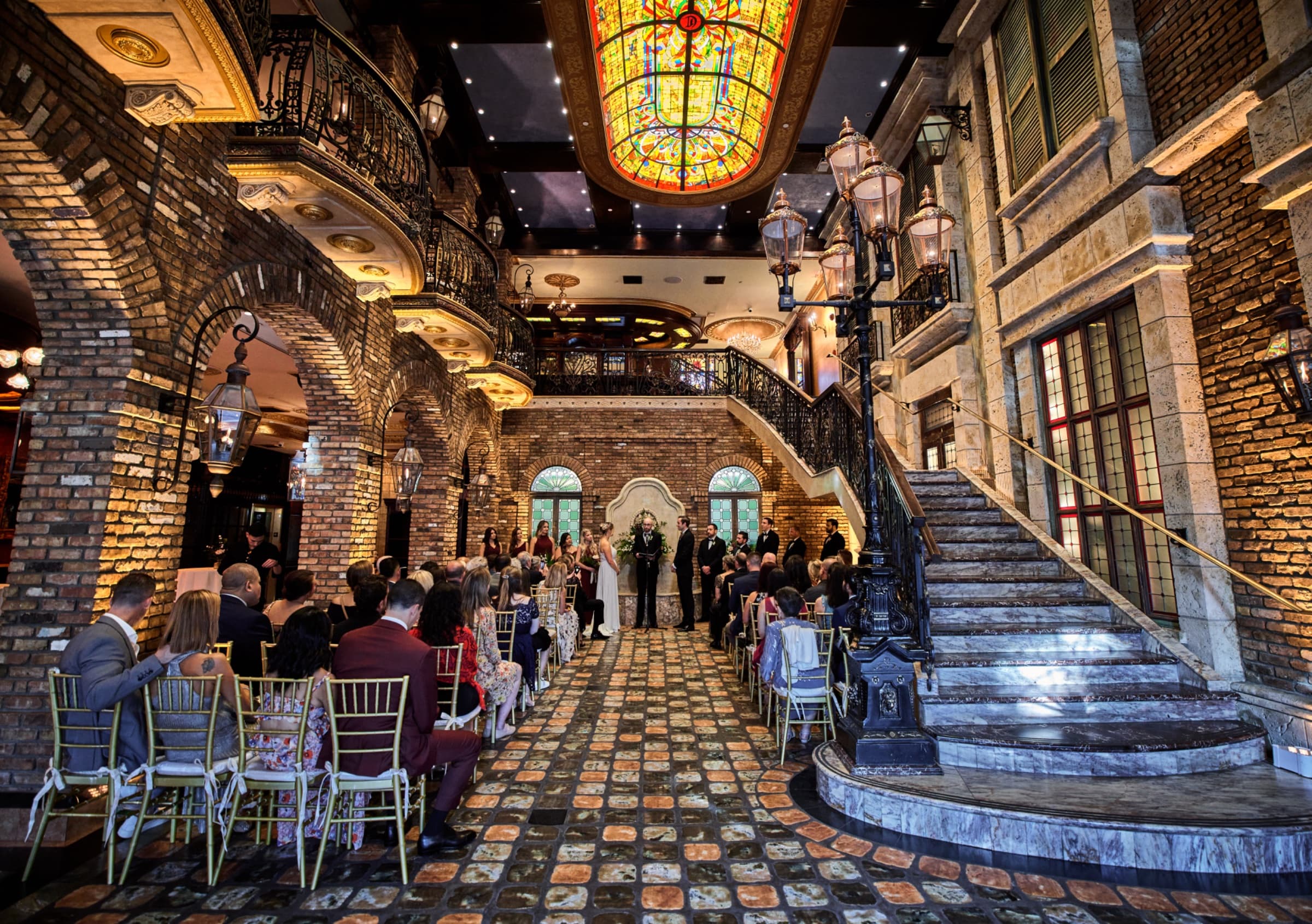 Ceremony beneath the stained-glass skylight with gold chiavari chairs and the wrought-iron staircase — The Cruz Building