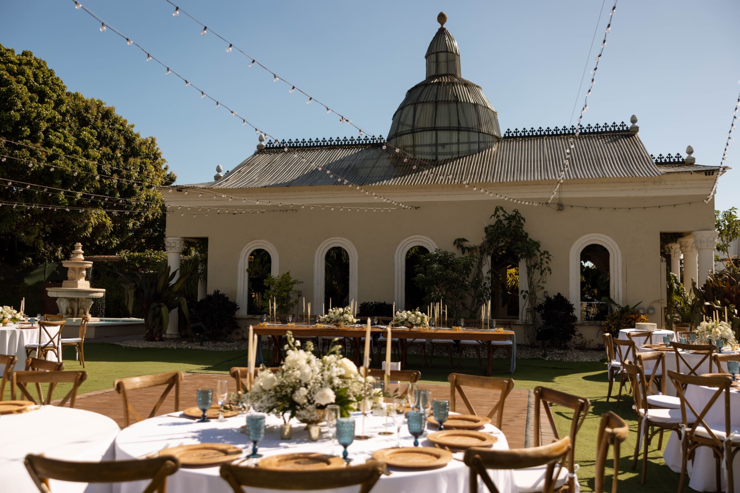 Outdoor courtyard reception under string lights with cross-back chairs, gold chargers and white floral centrepieces — Villa…